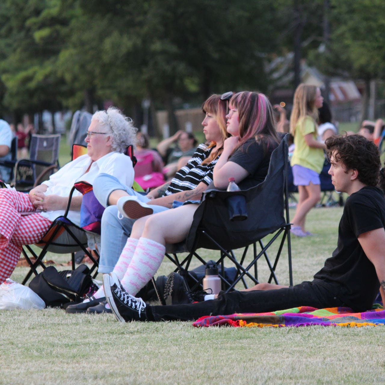 Families seated on lawn chairs and blankets on the grass at an outdoor concert in the park