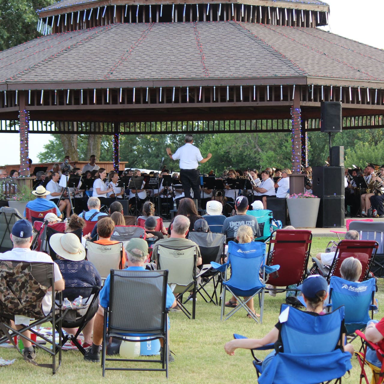 An orchestra performing under the park gazebo while an audience watches from lawn chairs